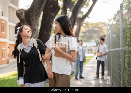 Two cheerful cute Asian female students friends are enjoying chatting while walking to school in the morning together, walking on a footpath, going ho Stock Photo