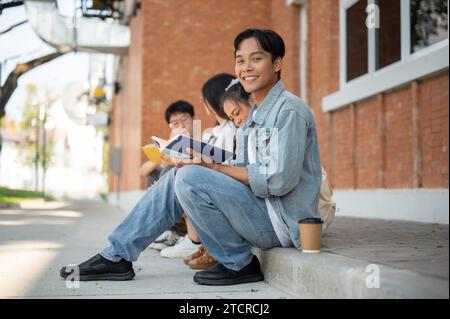 A happy young Asian man college student in jeans outfit sits on a footpath on his campus with his friends. Education and friendship concepts Stock Photo