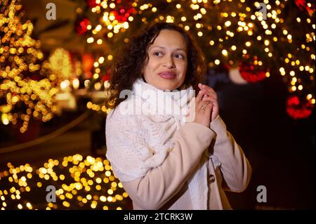 Smiling happy woman walking in Christmas market decorated with holiday lights in the evening. Feeling happy in big city. December. Christmas fairgroun Stock Photo