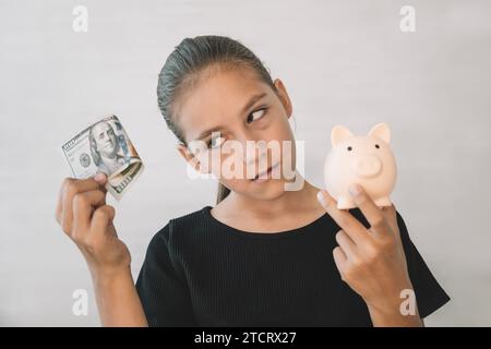 Portrait of delighted smiling little girl wearing black T-shirt standing and putting dollar banknote into piggy bank, saving money. Indoor on pink bac Stock Photo