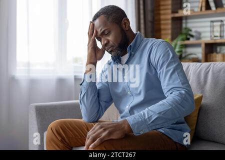Depressed mature senior gray haired man sitting sad alone by window on couch in living room, african american frustrated leaning on hand thinking about problems. Stock Photo