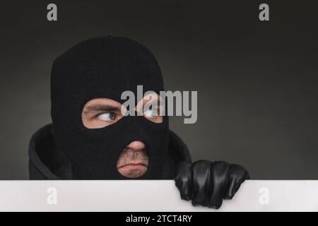 An evil thief in a mask looks out from behind a white board. Space for text and design. copy space. the surprised bandit looks away Stock Photo