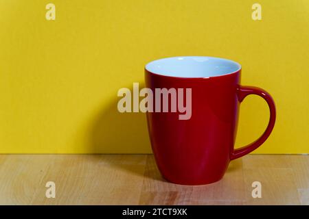 an empty red ceramic coffee mug with a handle stands on a natural wooden board in front of a colored background with real shadows Stock Photo