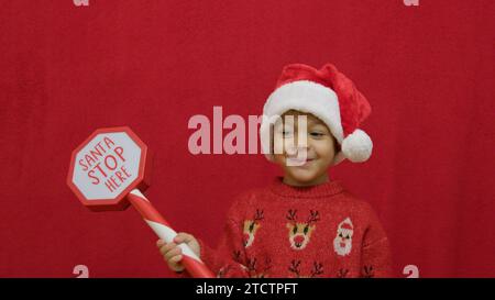 Cute preschool boy in christmas jumper waving sign Santa Stop Here while eating his cookies. Slow motion isolated on red. High quality photo Stock Photo