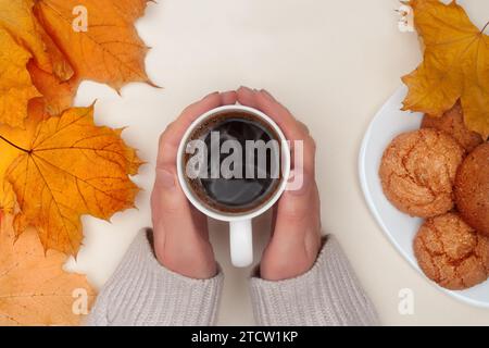 Flat lay of autumn yellow maple leafs, woman's hands hold cup of hot tea and white plate with cookies on ivory background. Top view. Stock Photo