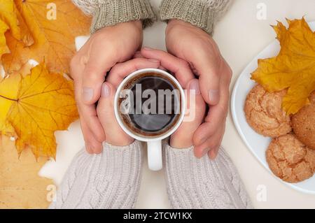 Top view of autumn yellow maple leafs, man's hands over woman's hands waht hold cup of hot coffee, white plate with cookies on ivory background. Cozy Stock Photo