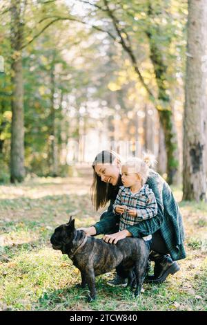 Little girl stands next to her mother stroking a black french bulldog in the forest Stock Photo