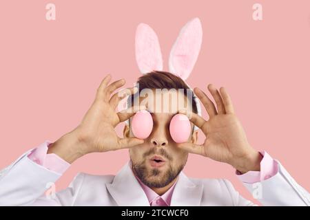 Funny surprised young man wearing cute bunny ears covering his eyes with two Easter eggs Stock Photo