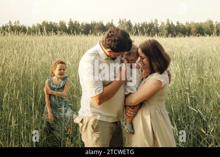 Dad and mom are holding their little daughter in their arms, hugging her, with their eldest daughter nearby. Standing in a field of green wheat at sun Stock Photo