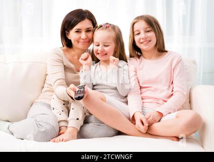 Portrait of mom hugging her cute daughters while sitting on the sofa and watching TV at home. Happy family relaxing on the coach Stock Photo