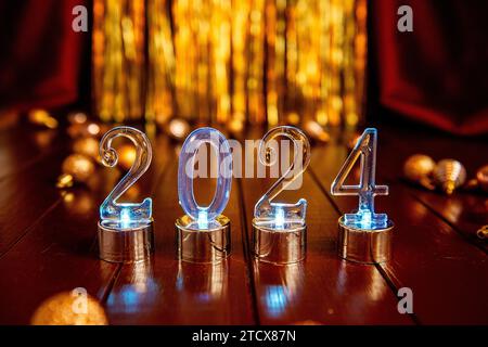 On wooden brown background transparent blue numbers 2024 among Christmas decorations balls on texture of foil golden tinsel, burgundy fabric. New Year Stock Photo