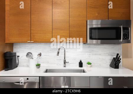 A kitchen faucet detail with wood and stainless steel cabinets, a white marble tiled backsplash, and kitchen utensils on the marble countertop. Stock Photo