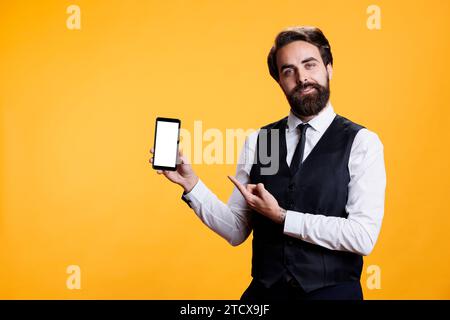 Professional employee advertises white screen on mobile phone, showcasing empty isolated blank display with copyspace against yellow background. Young man presents blank mockup device. Stock Photo