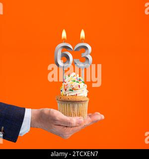 Hand holding birthday cupcake with number 63 candle - background orange Stock Photo