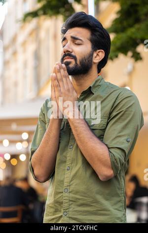 Bearded religion indian man praying with closed eyes to God asking for blessing, help, forgiveness outdoor. Adult guy clasping hands wishing luck in urban sunny city street. Town lifestyle. Vertical Stock Photo
