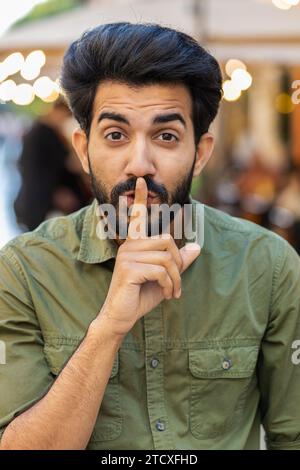 Shh be quiet please. Indian man presses index finger to lips makes silence hush gesture sign do not tells gossip secretoutdoors. Arabian guy walking in urban city street. Town lifestyles. Vertical Stock Photo