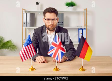 Pensive young man sitting at desk in office looking at flags of different countries Stock Photo