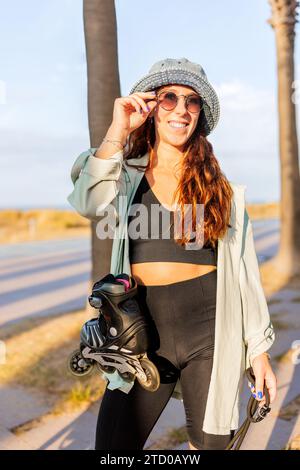 Happy young beautiful Caucasian brunette woman wearing hat and sunglasses standing with inline roller skates against sky in park Stock Photo
