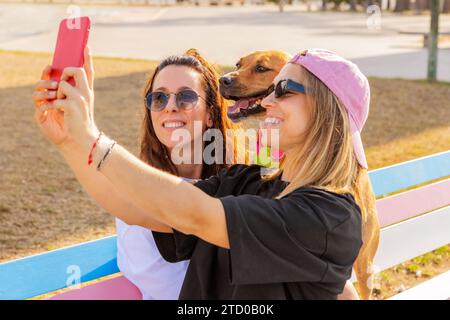 Cheerful Caucasian young woman taking selfie with female friend and dog through smartphone while sitting together at park during weekend Stock Photo