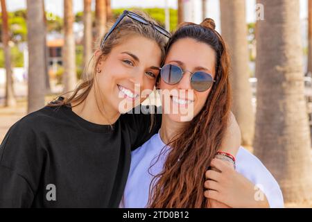 Portrait of happy Caucasian young blond woman with arm around brunette friend wearing sunglasses at park during weekend Stock Photo