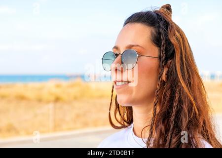 Portrait of smiling attractive young woman with long brown hair and braids wearing sunglasses and looking away against clear sky Stock Photo