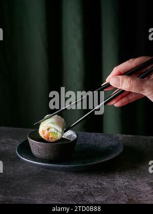 Crop hand of anonymous person using chopsticks to elegantly hold a sushi roll above a small bowl of soy sauce against dark green curtains Stock Photo