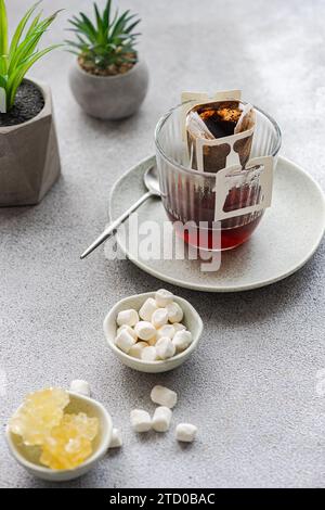A fresh cup of drip coffee brewing in a clear glass with trendy grey backdrop, accompanied by sweet marshmallows and chic decor plants. Stock Photo