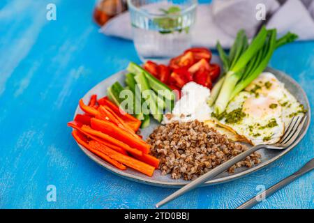 A nutritious breakfast setup showcasing sliced raw vegetables, feta cheese, buckwheat, and fried eggs with pesto on a blue background. Stock Photo