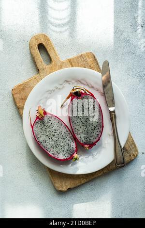 A halved dragonfruit with vibrant pink skin and speckled flesh presented on a wooden serving board with a knife. Stock Photo