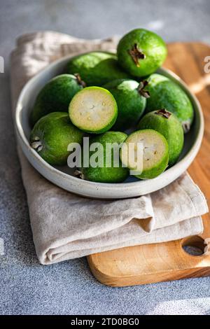Raw feijoa fruits, also known as pineapple guavas, displayed in a ceramic bowl on a wooden cutting board with a linen cloth. Stock Photo
