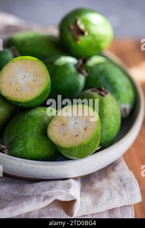 A close-up view of fresh feijoa fruits, with one sliced in half, in a ceramic bowl on a linen cloth. Stock Photo