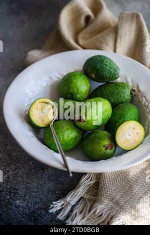 A composition of ripe feijoa fruits arranged in a white ceramic bowl, one cut open, with a textured napkin on a dark surface. Stock Photo