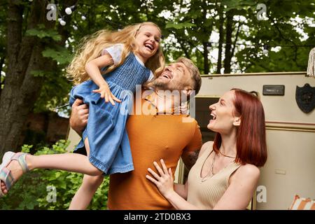 father holding  overjoyed daughter near smiling wife and modern trailer home, fun and laughter Stock Photo