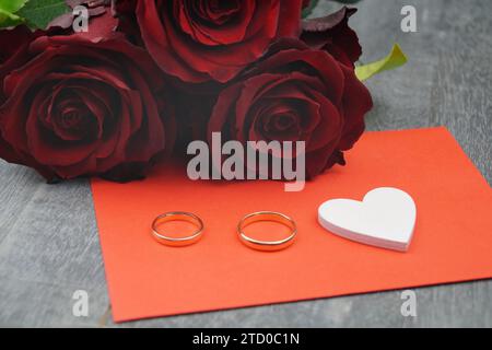 hybrid tea (Rosa odorata hybrida), wedding rings in front of baccarat roses next to a white wooden heart on a red envelope Stock Photo