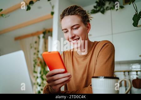 Low angle of Smiling young female in casual clothes and with dreadlocks looking at screen of mobile phone while sitting at kitchen table with laptop c Stock Photo