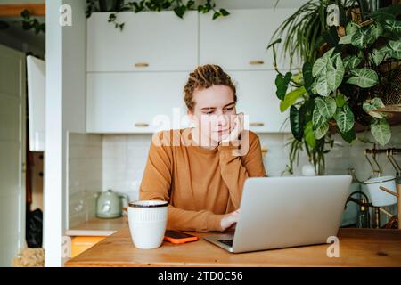 Thoughtful young female with dreadlocks looking at screen of laptop and reading recipe while sitting leaning chin on hand at kitchen table with mobile Stock Photo