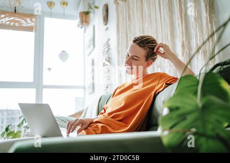 Young female freelancer smiling and working on project while sitting on comfortable sofa with cushions at home near green plants Stock Photo