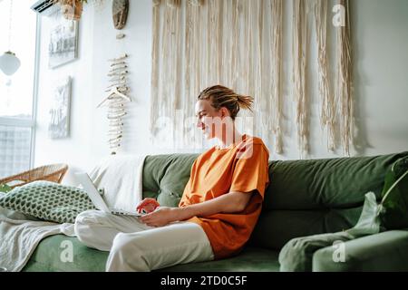 Side view of Young female freelancer smiling and working on project while sitting on comfortable sofa with cushions at home near window Stock Photo