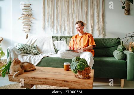 Young female freelancer smiling and working on project while sitting on comfortable sofa with cushions at home near adorable British shorthair golden Stock Photo