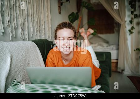 Happy young woman lying on comfortable sofa and browsing laptop while working online at home Stock Photo
