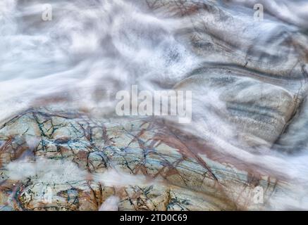 Smooth water flows create a ghostly veil over the multi-colored stone surface beneath. Stock Photo