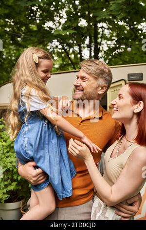 father holding cheerful daughter near pleased wife and modern trailer home, leisure and adventure Stock Photo