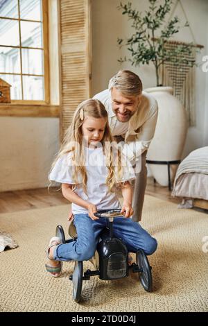 smiling man assisting cute daughter riding toy car in modern living room at home, playing together Stock Photo