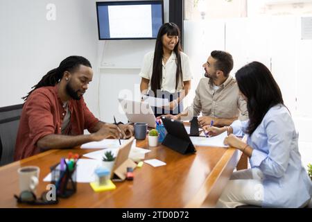 Serious business multiethnic people examining documents and using tablet while colleagues discussing over laptop in background during meeting in offic Stock Photo