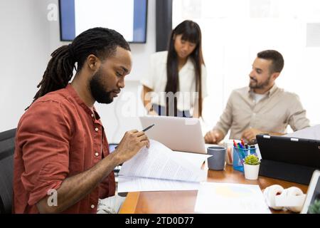 Serious busines black person examining documents while colleagues discussing over laptop in background during meeting in office Stock Photo