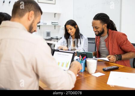 Happy business people examining documents and using tablet while other anonymous colleagues work on the computer background during meeting in office Stock Photo