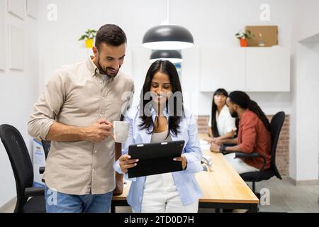 Smiling multiracial man holding coffee mug and discussing with female colleague over digital tablet while standing in boardroom at creative office Stock Photo