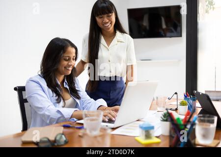 Smiling female manager looking at businesswoman working on laptop while sitting at desk in creative office Stock Photo