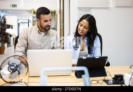 Smiling multiracial male and female colleagues discussing plan over digital tablet and laptop while sitting at desk in creative office Stock Photo