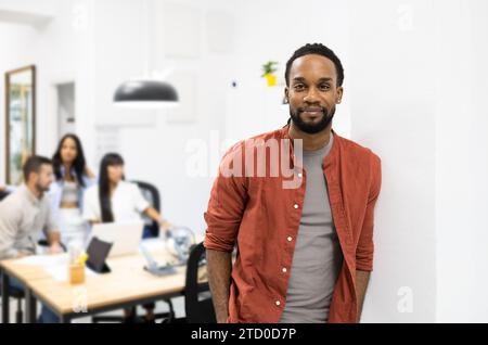 Portrait of confident African American young businessman wearing casuals standing against wall and looking at camera in creative office Stock Photo